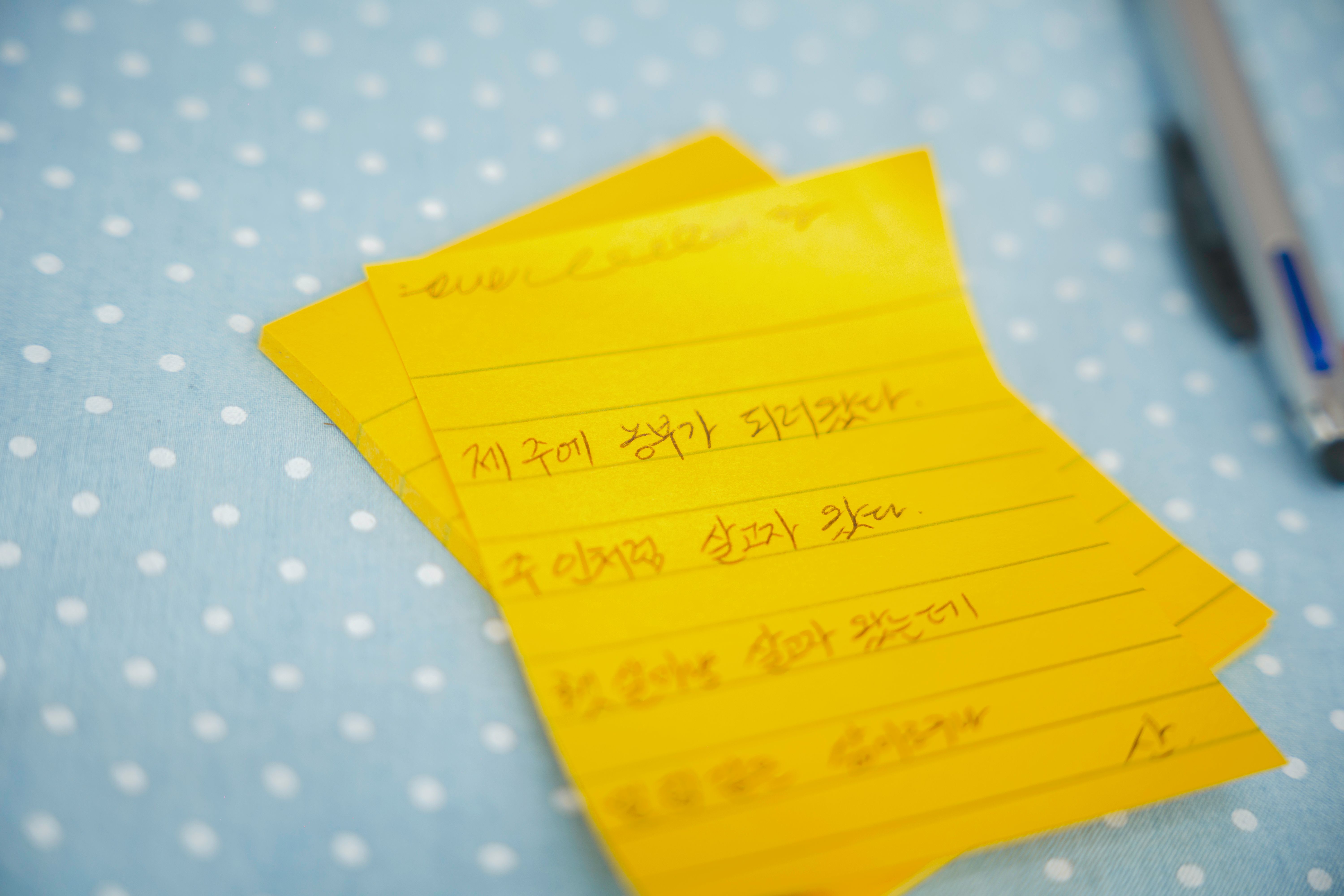 A stack of yellow lined sticky notes containing handwritten Korean text sits on a light blue polka-dotted surface next to a silver pen
