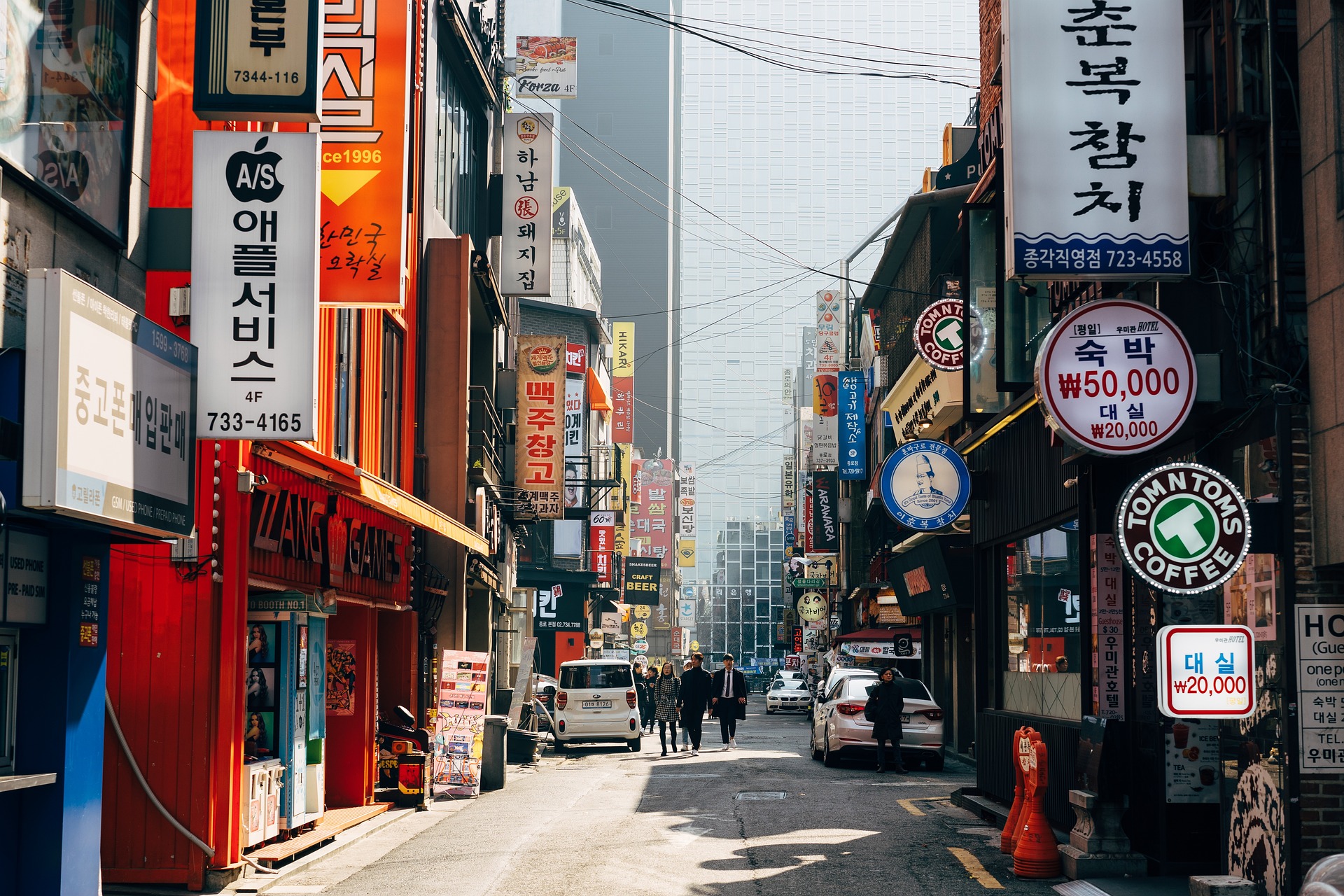 Busy Korean shopping street lined with colorful Hangul signs, cafes, shops, cars, and pedestrians under a web of overhead utility wires