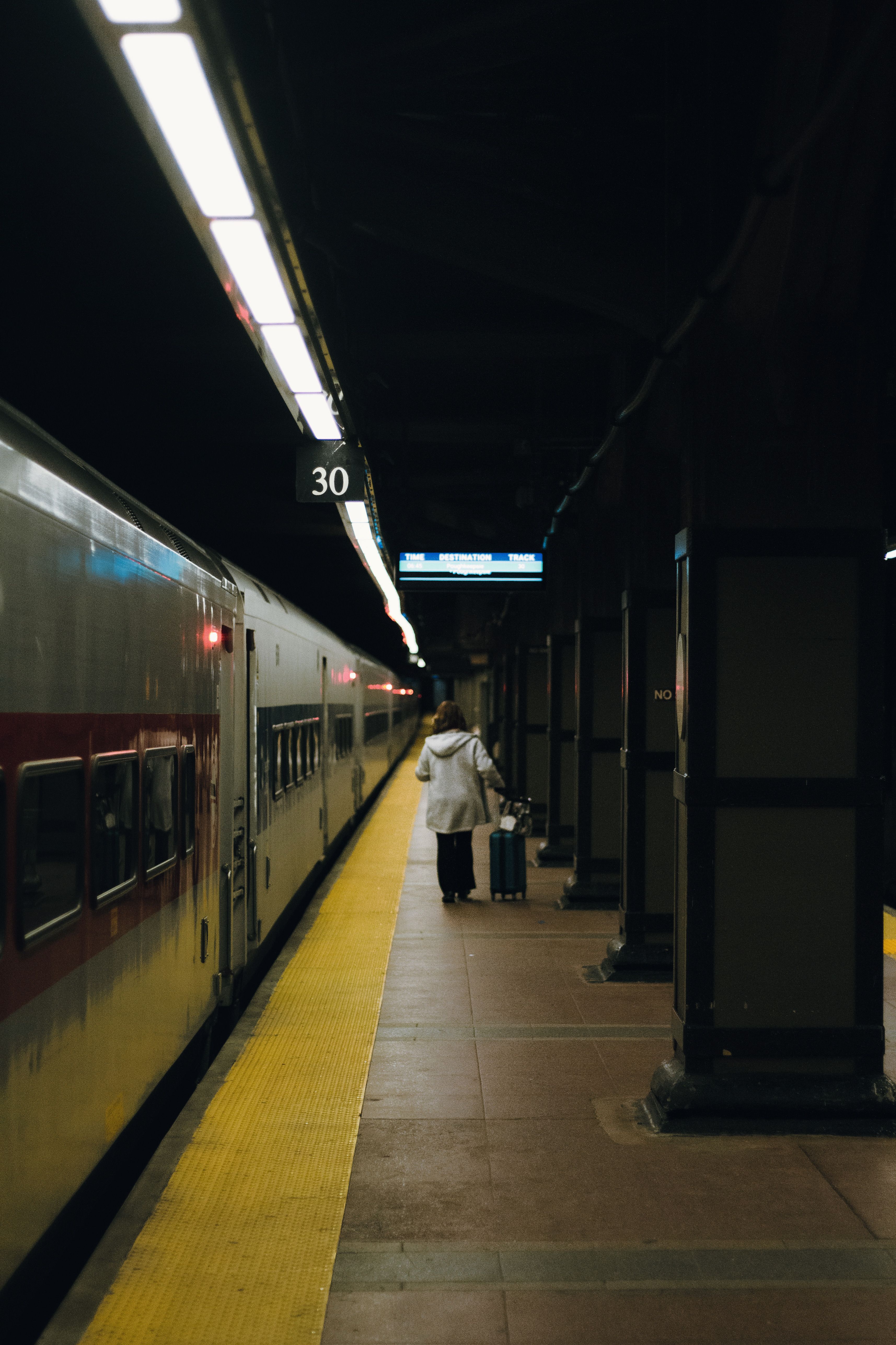 A person pulling a wheeled suitcase walks away along a dimly lit underground train platform next to a stationary passenger train
